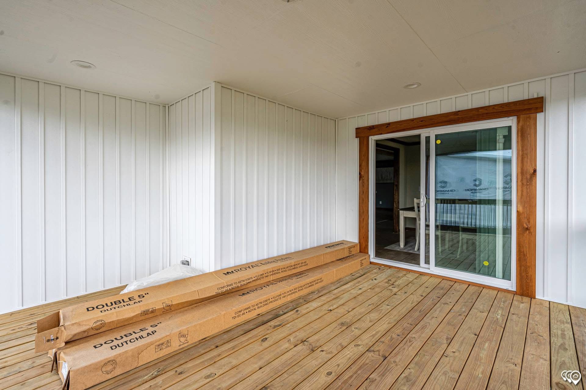 A newly constructed porch with light wood flooring and white paneled walls. Two long cardboard boxes are on the floor near a glass sliding door framed with dark wood. The atmosphere is clean and modern.