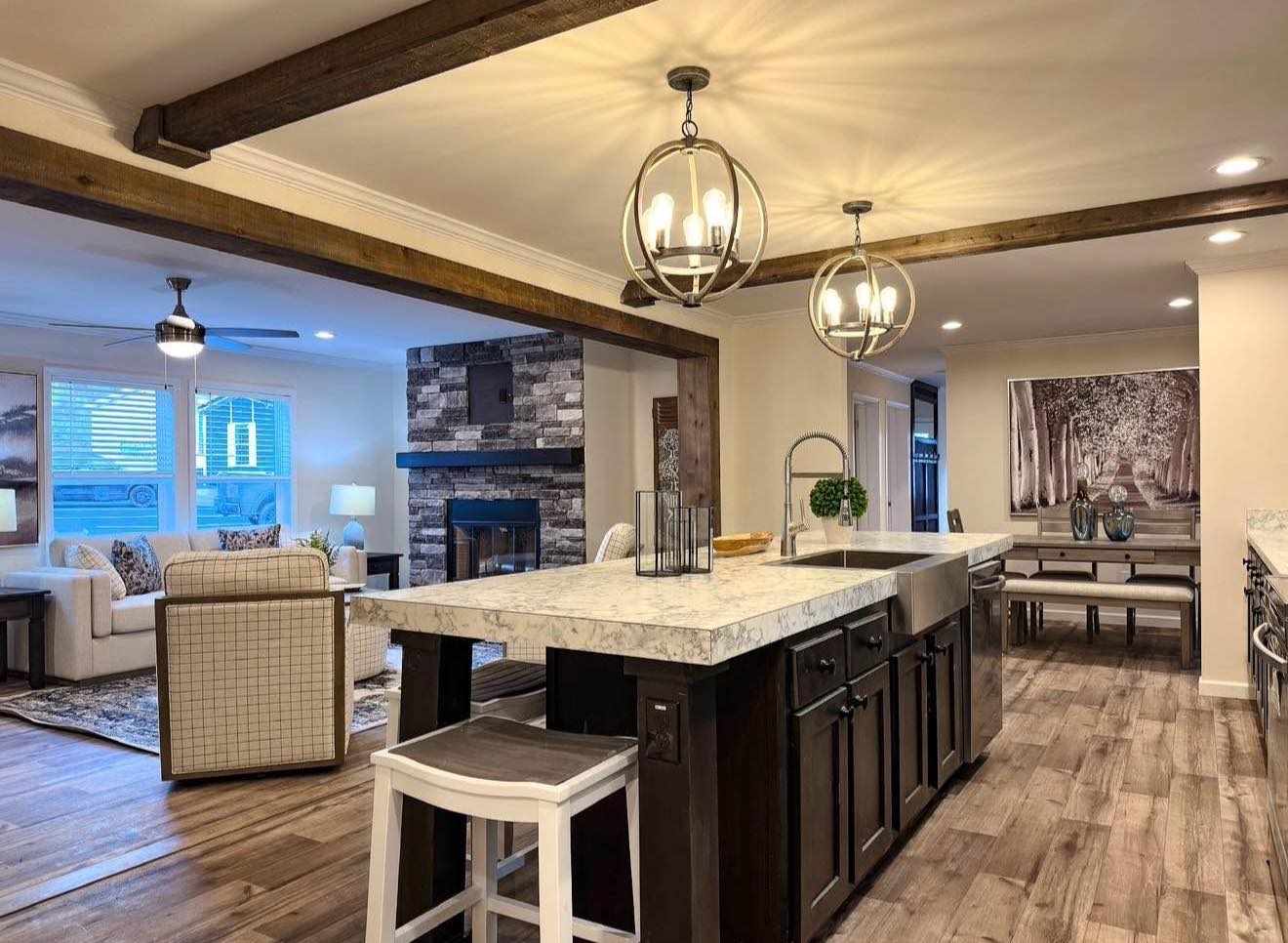 Spacious open kitchen and living area with wooden beams, marble countertops, and warm lighting. Cozy stone fireplace and artful wall hangings. Calm ambiance.