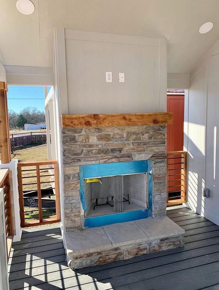 Outdoor stone fireplace under construction on a wooden deck. Blue tape covers the opening, and sunlight casts shadows on the deck, creating a calm atmosphere.