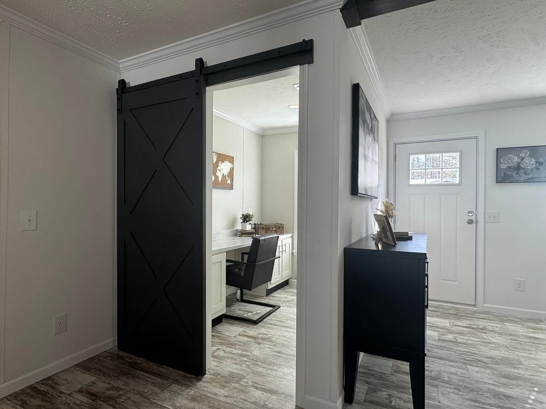 Cozy room with a white and gray color scheme. Features a dark sliding barn door, home office, decorative artwork, and sunlight through the front door.