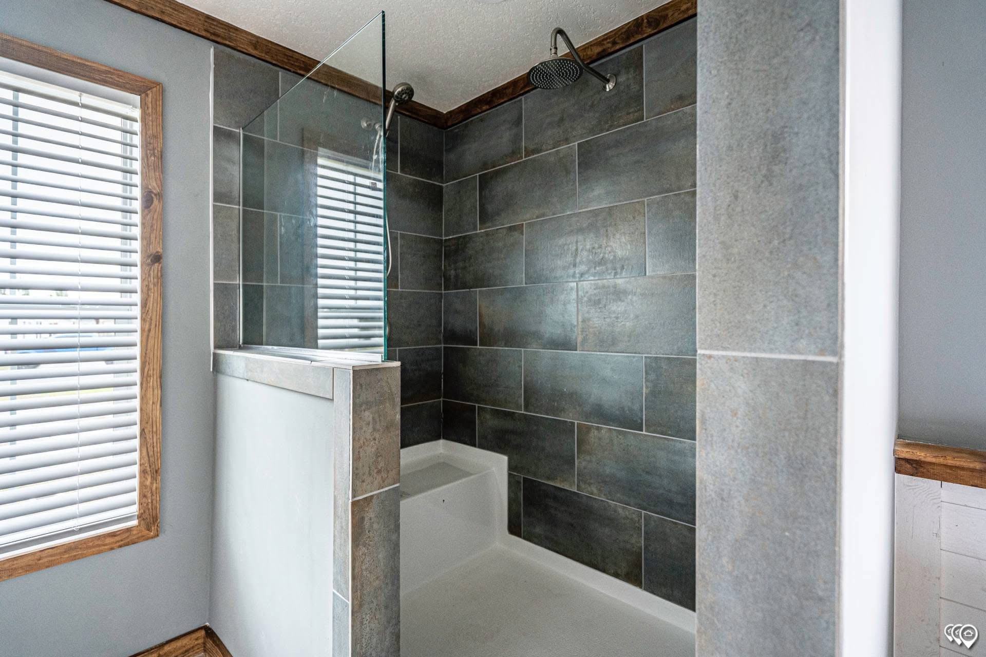 Modern bathroom featuring a large walk-in shower with dark gray tiled walls, a rain showerhead, and a glass partition. Bright window with blinds.