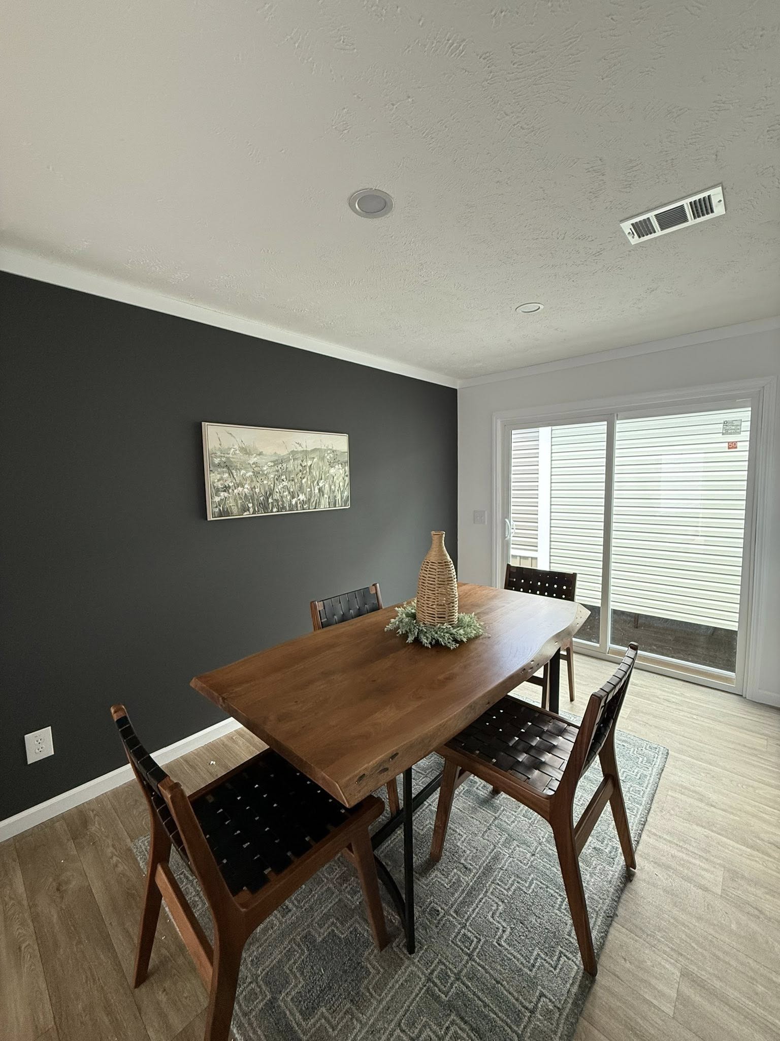 Modern dining room with a wooden table and four chairs on a patterned rug, set against a dark accent wall. A vase with greens decorates the table, and sliding glass doors allow natural light.
