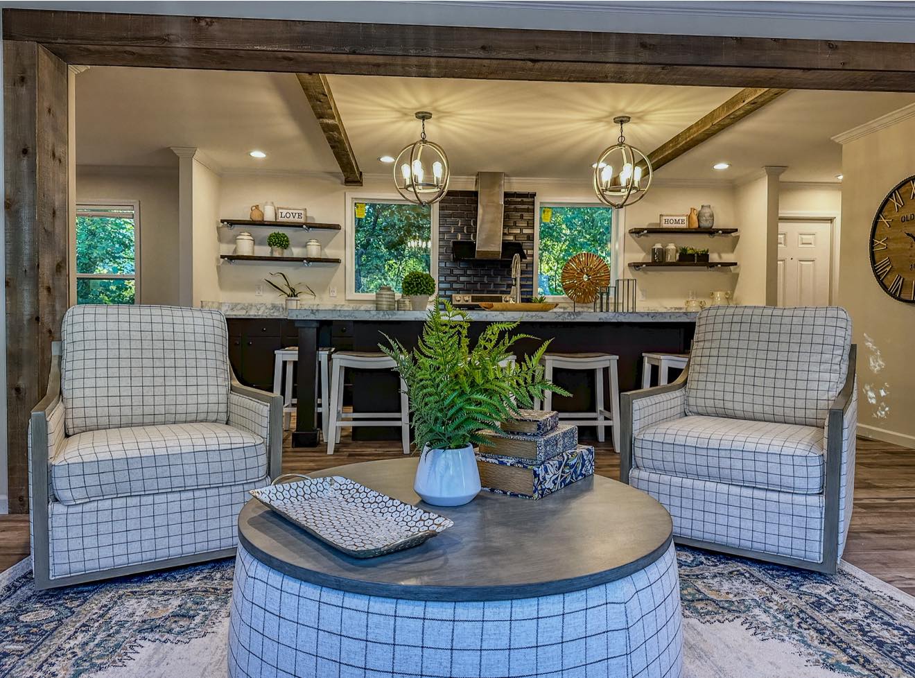 Cozy living room with two plaid armchairs and a round coffee table featuring a fern and stacked books. Open kitchen with hanging lights in the background.