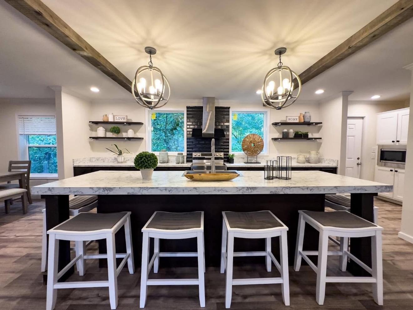Modern kitchen with marble island, white stools, and orb chandeliers. Two large windows, open shelves, black backsplash, and wooden accents create a cozy, elegant feel.