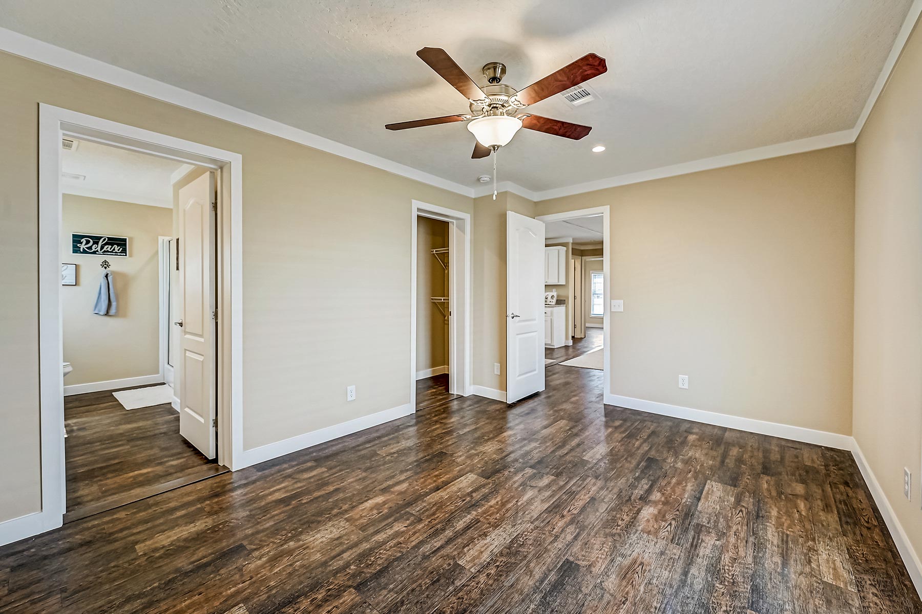 Empty room with tan walls and dark wood flooring. Features a ceiling fan with light, open doors to a closet, bathroom, and a kitchen in the background.
