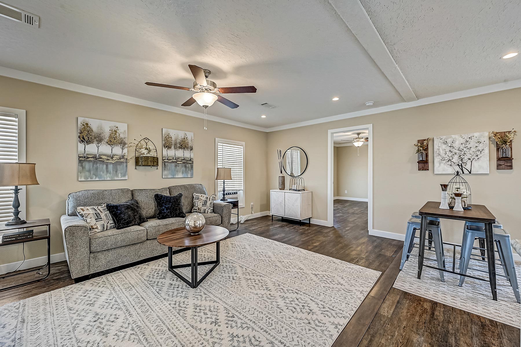 A cozy living room with a gray sofa, patterned cushions, and a fur throw. A round wooden coffee table sits on a large, patterned rug. Beige walls display nature art, and a ceiling fan hangs above. A dining area with a table and metal chairs is visible. The atmosphere is warm and inviting.