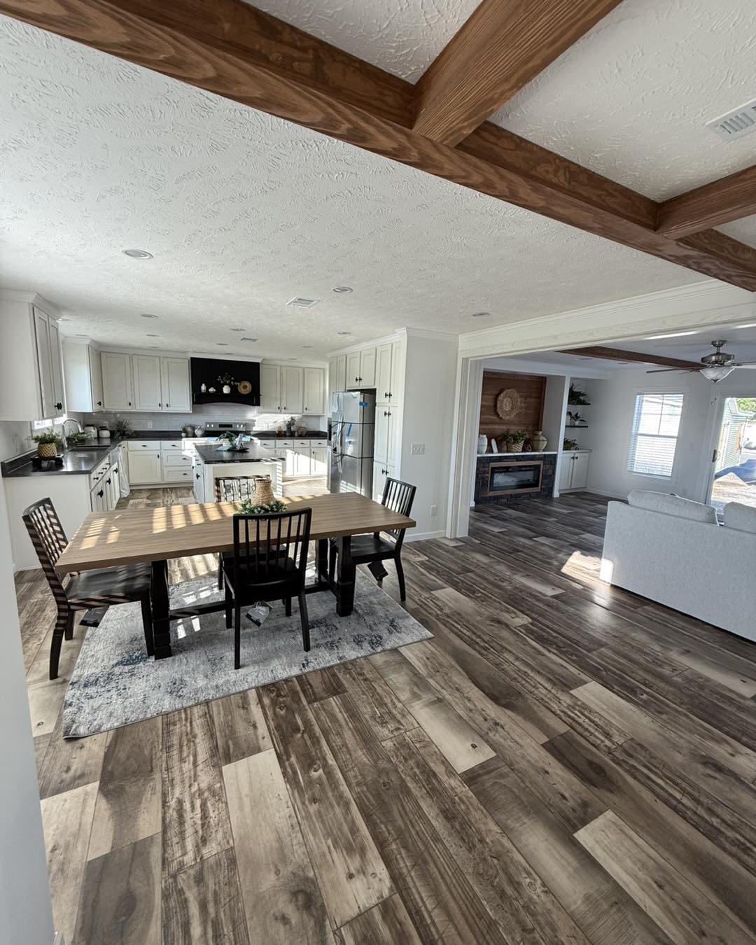Spacious kitchen with white cabinets, black countertops, and wood beams. A dining table with dark chairs sits on wood flooring, leading to a cozy living area.