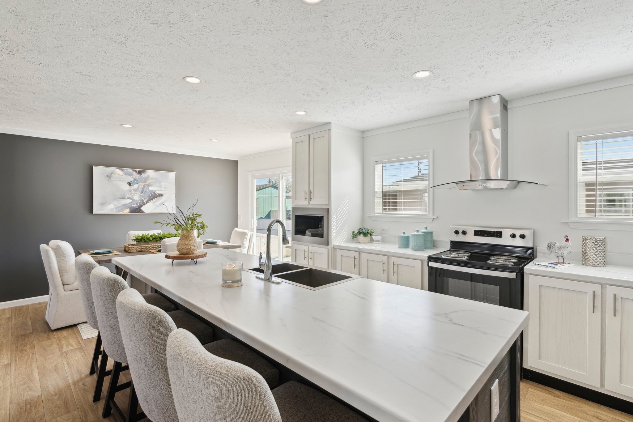 Modern kitchen with white cabinets and a large marble island, featuring cushioned chairs. Sunlit room with wooden floor, sleek appliances, and wall art.