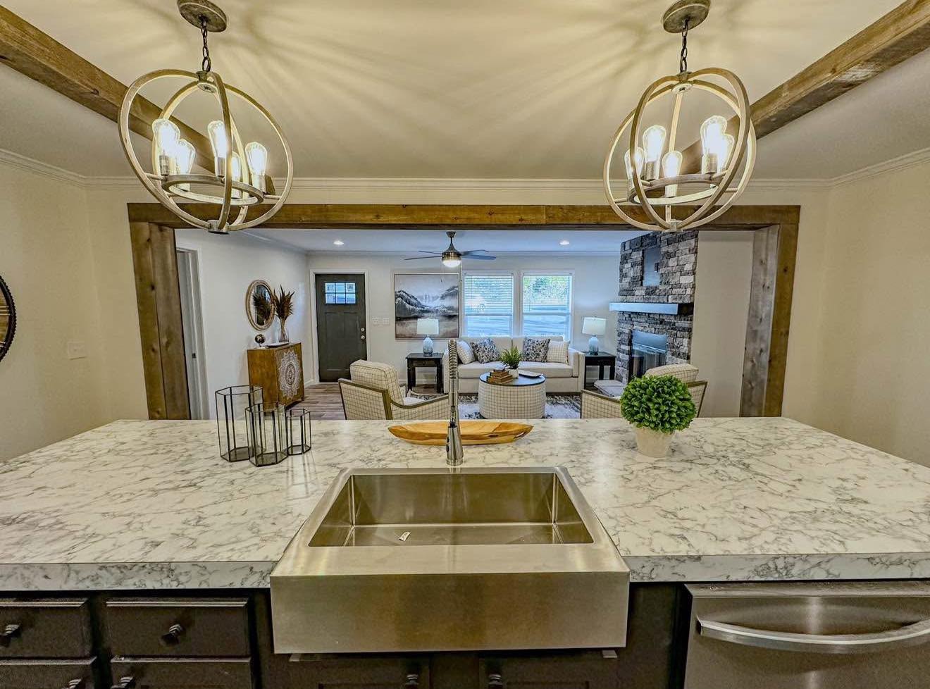 Modern kitchen view with a marble counter and stainless steel sink. Beyond, a cozy living room with a stone fireplace, neutral tones, and warm lighting.