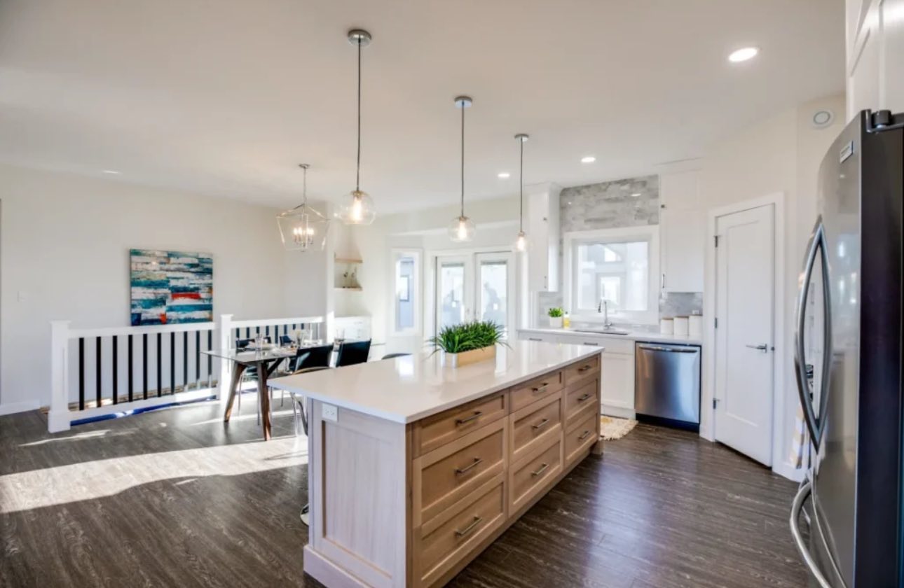 Bright, modern kitchen with a large island featuring light wood drawers, pendant lighting, and stainless steel appliances. Dining area and colorful art add warmth.