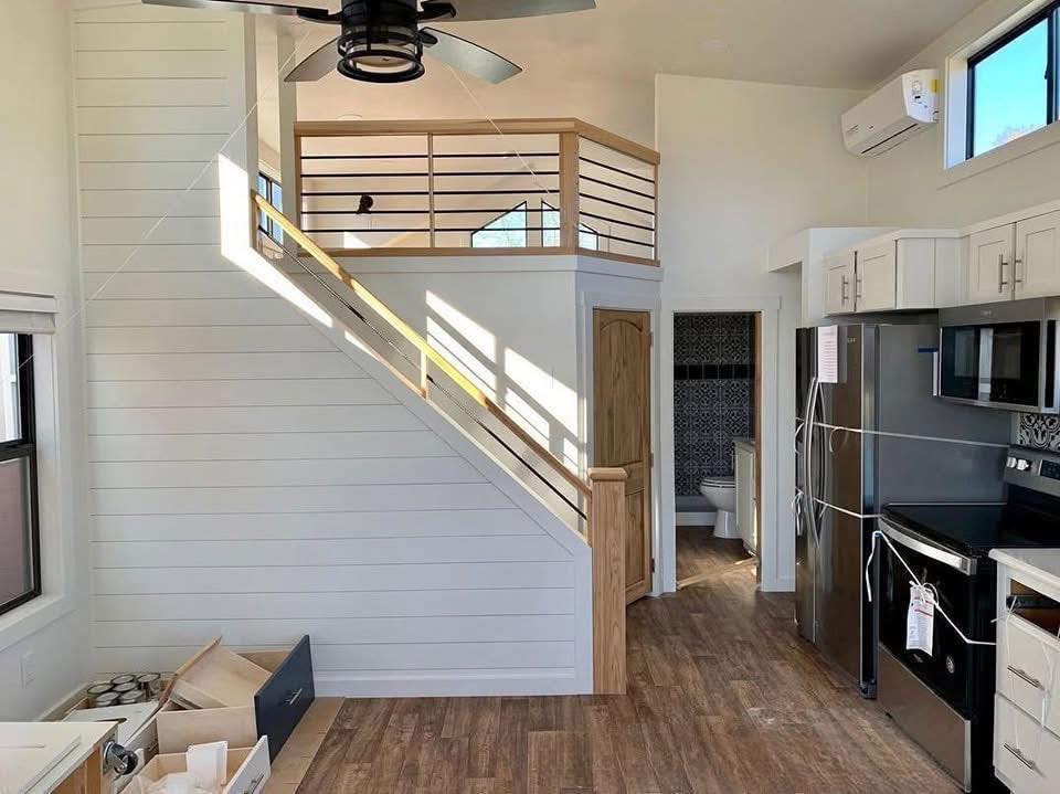 Modern interior of a tiny house featuring a loft with wooden railings, white shiplap walls, and wood flooring. A kitchen and staircase lead to a cozy upper area.