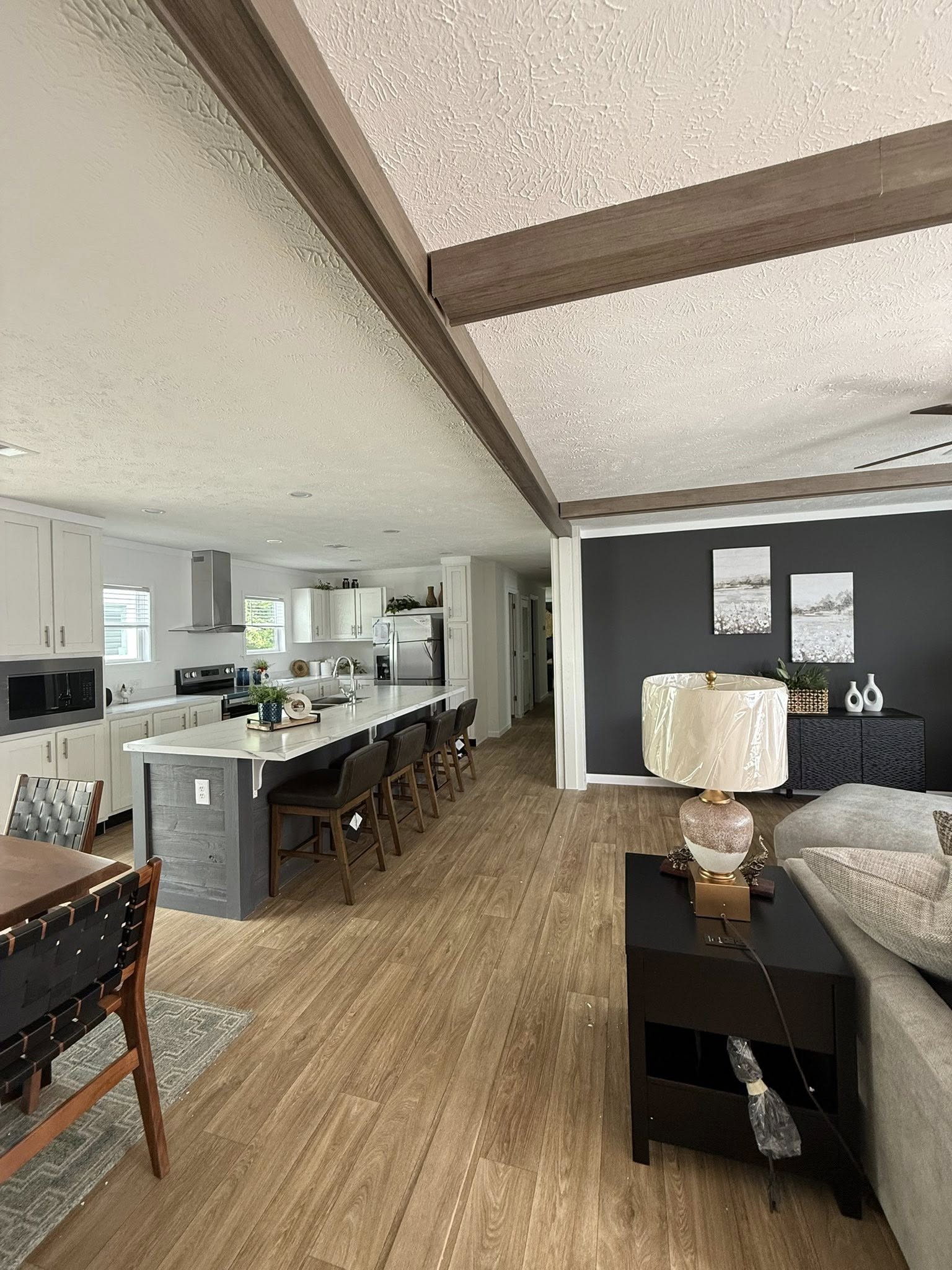 Open-concept living space with light wood flooring. Kitchen features a white island and black chairs. Cozy seating area has a lamp and dark table. Neutral tones create a calm ambiance.