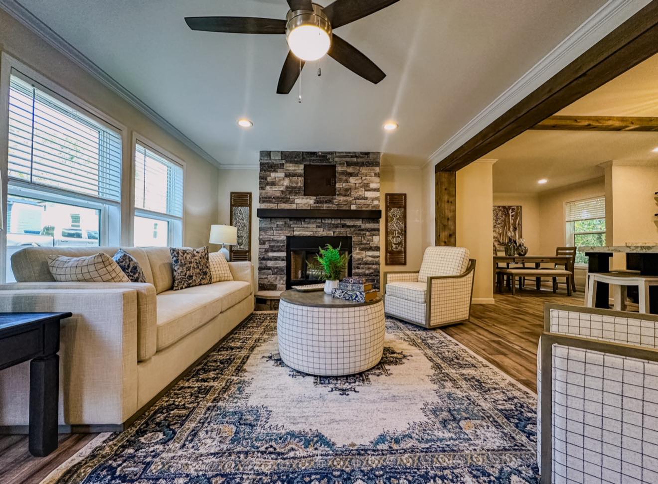 Warm living room with beige sofa, plaid chairs, and a round ottoman on a blue-patterned rug. Stone fireplace and ceiling fan enhance coziness.