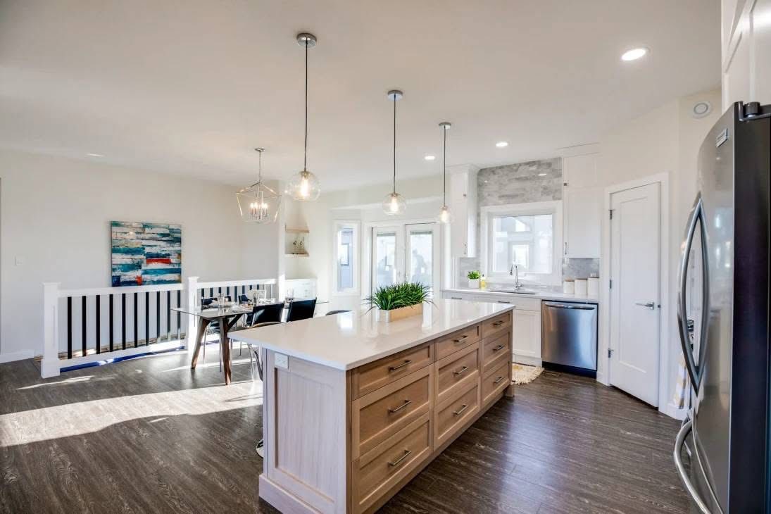 Modern kitchen with a large island featuring light wood cabinets and white countertop. Pendant lights hang above. Dining table and abstract art seen in the background.