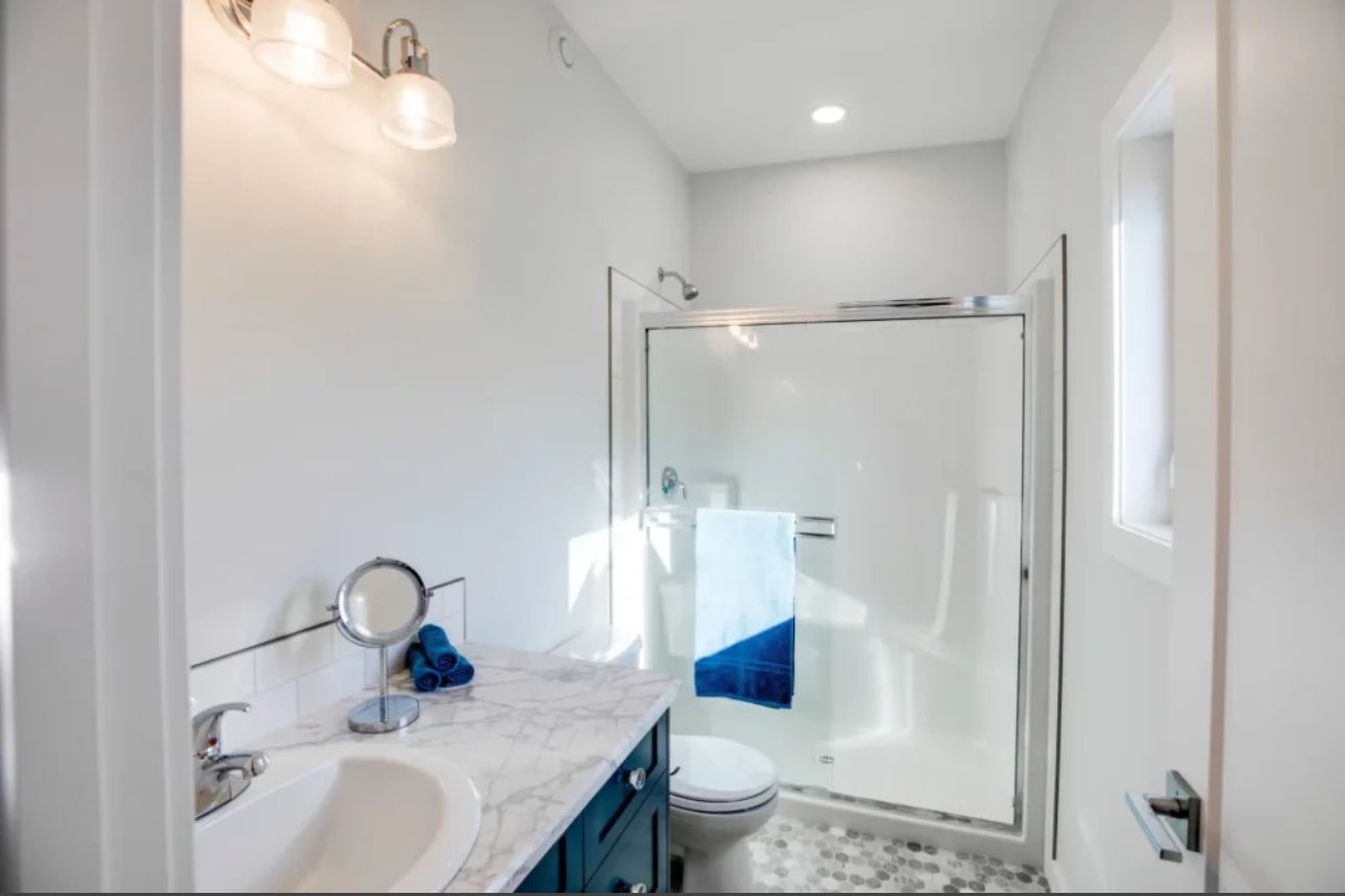Bright bathroom with white walls and hexagon tile flooring. Features a glass shower, marble countertop, blue vanity, and modern lighting, creating a clean, fresh atmosphere.