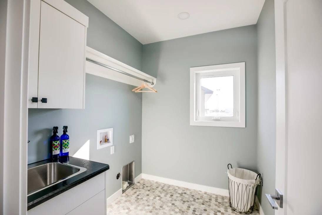 Laundry room with light gray walls and a window. Features include a black countertop with a sink, hanging rail, and laundry basket. Tidy and bright.