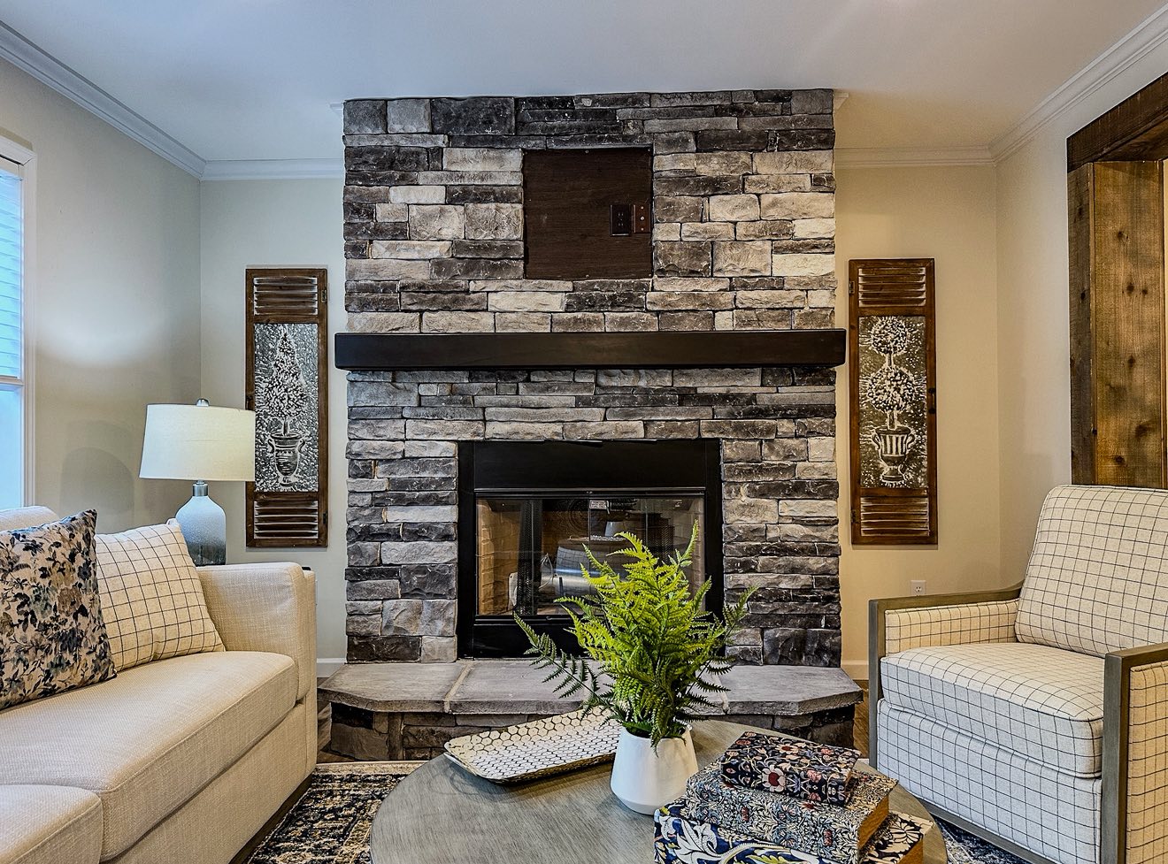 Cozy living room with a stone fireplace centered between two art pieces. Cream sofa and plaid armchair face a round table with a plant, exuding warmth.