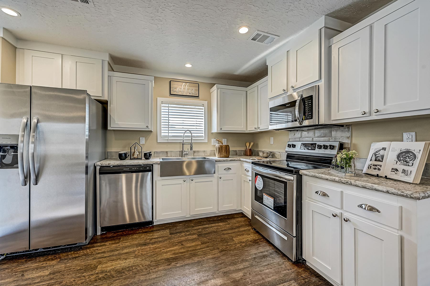 Modern kitchen with white cabinets, stainless steel appliances, and wooden floors. A central window casts soft light, creating a cozy atmosphere.