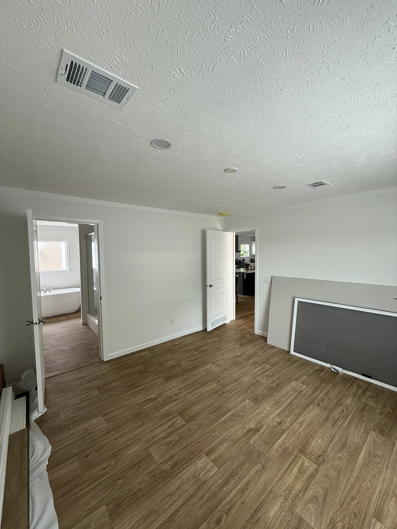 Empty room with wood flooring and white walls, featuring two open doors leading to a bathroom and a kitchen. A dismantled piece of furniture leans against the wall, conveying a sense of ongoing renovation or move.