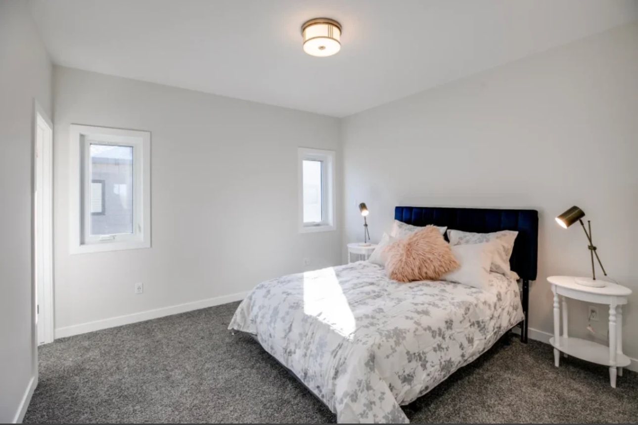 A bright bedroom with white walls, gray carpet, and a bed with floral bedding and pink cushions. Two side tables with lamps add a cozy feel.