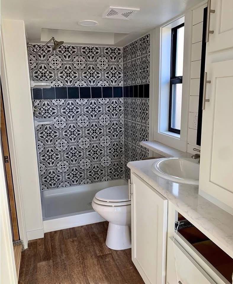 Compact bathroom with patterned black-and-white tiles in the shower, a white toilet, wooden floor, white vanity with sink, and bright light from a window.