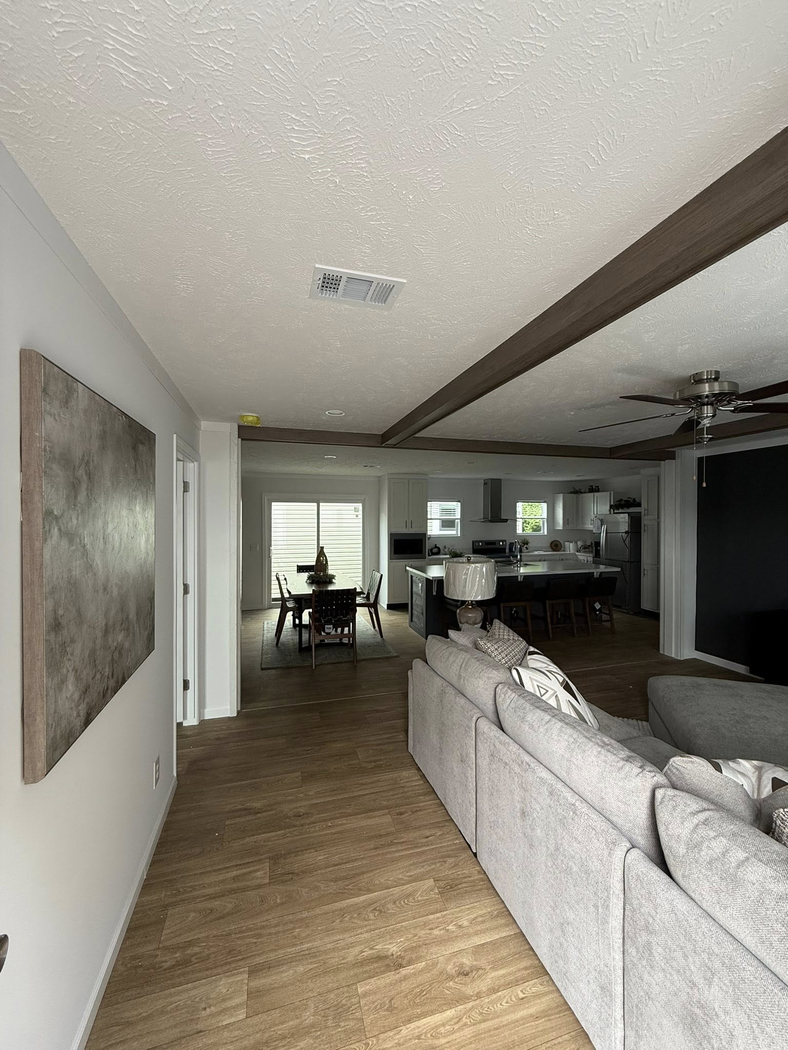 Modern living area with wood floors, a gray sectional sofa, and an open kitchen. Dining table at the back near glass doors, creating a cozy atmosphere.