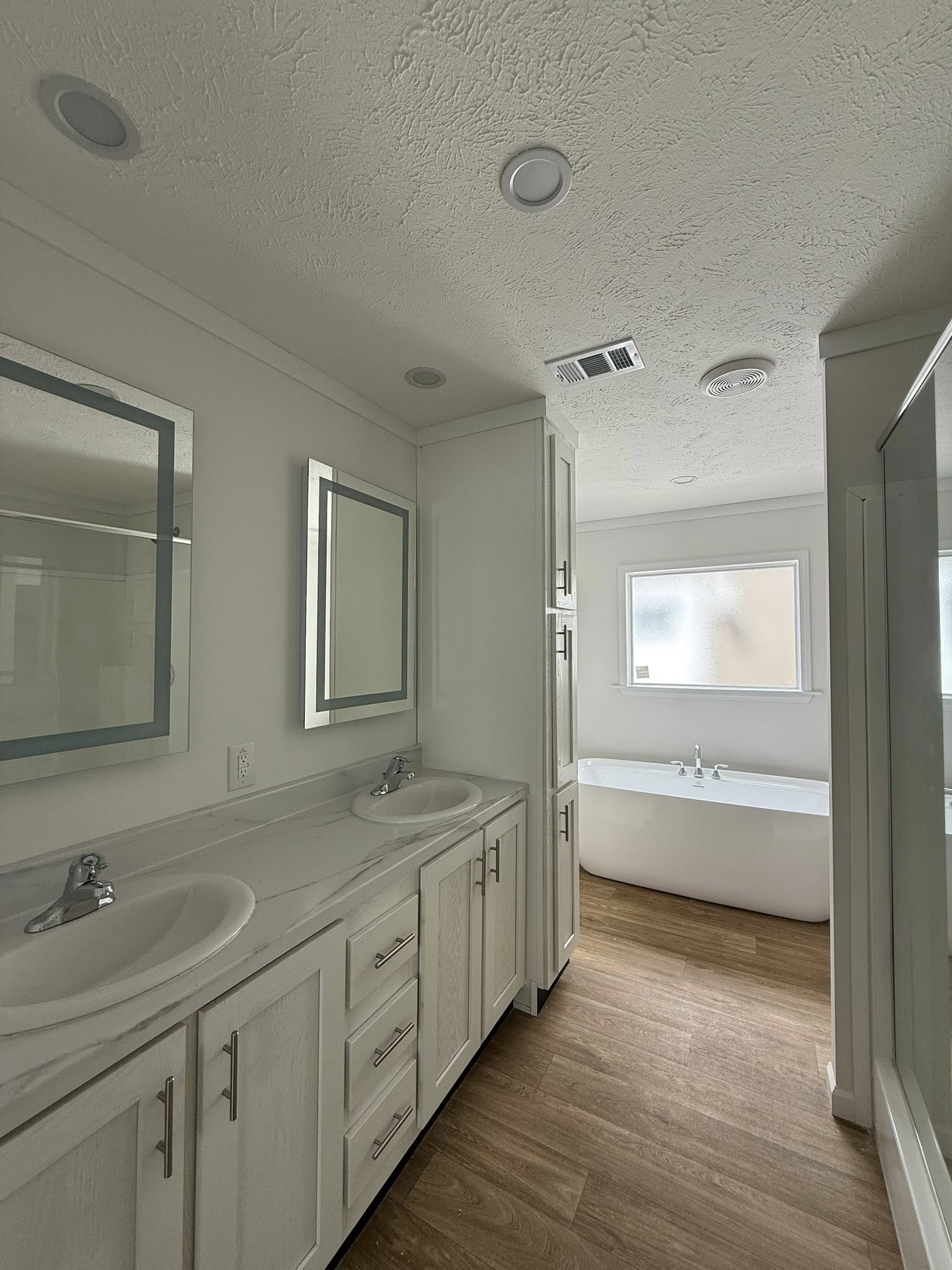 Bright bathroom interior with dual sinks and mirrors, white cabinetry, and a freestanding bathtub near a frosted window. The wooden floor adds warmth.
