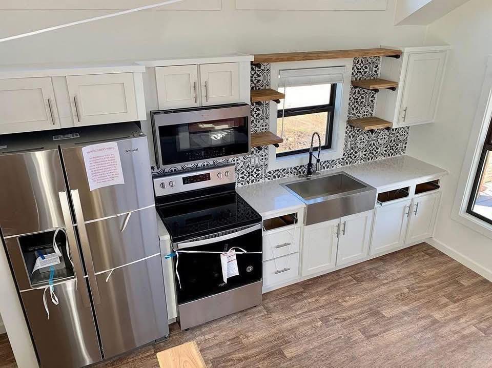 Modern kitchen with light wood floors features a stainless steel fridge, oven, and farmhouse sink. White cabinets, open shelves, and patterned tile add elegance.