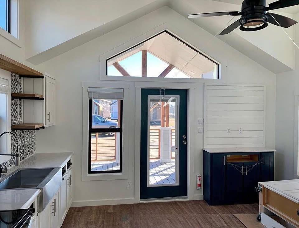 A bright, modern kitchen features white cabinets and patterned tile backsplash on the left. A glass-paneled door leads to a sunny porch, with a high ceiling and a black ceiling fan.