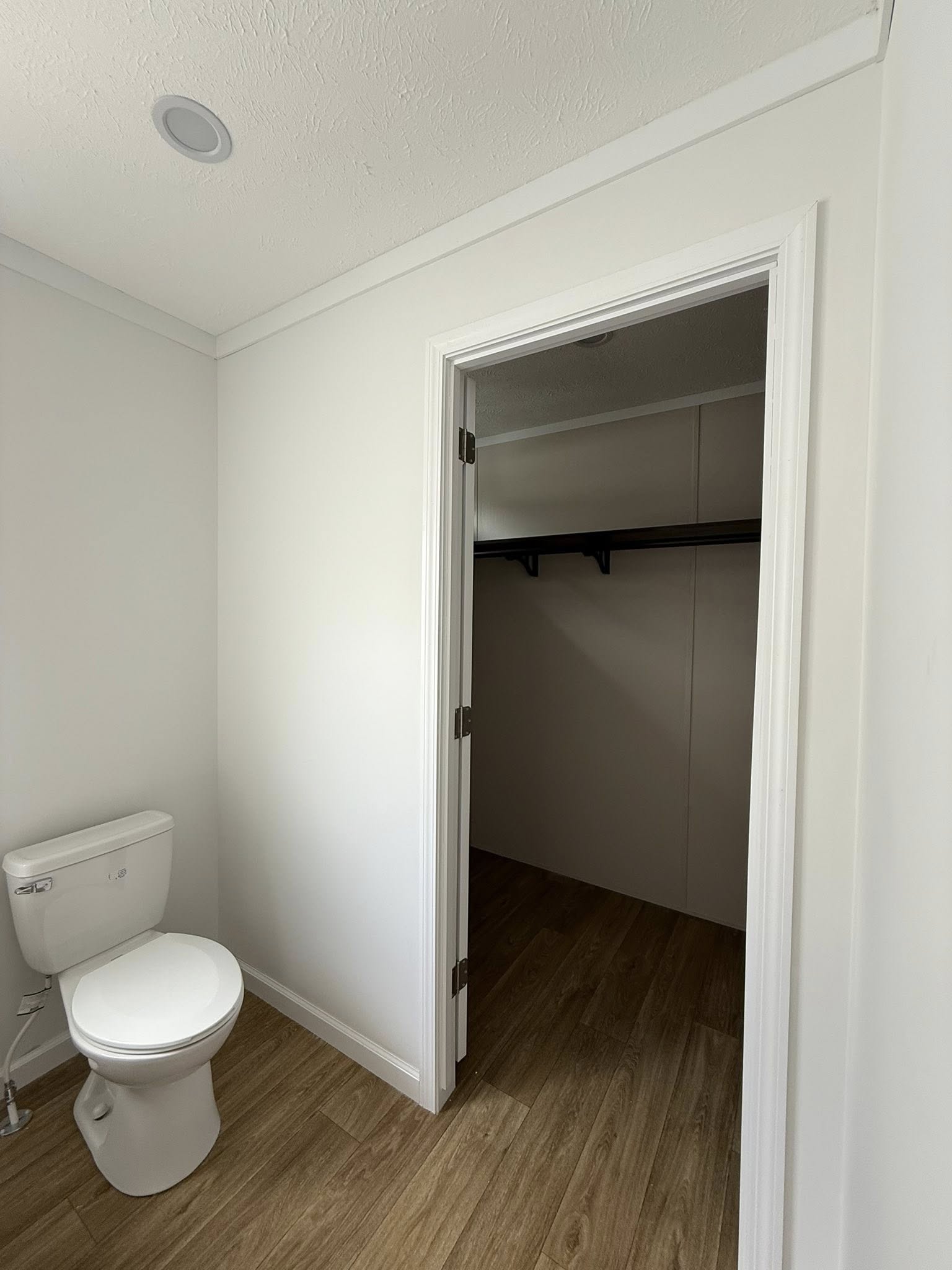 A simple, white bathroom features a toilet with wood flooring. An open door reveals a walk-in closet with a wooden shelf, creating a clean, minimalist feel.