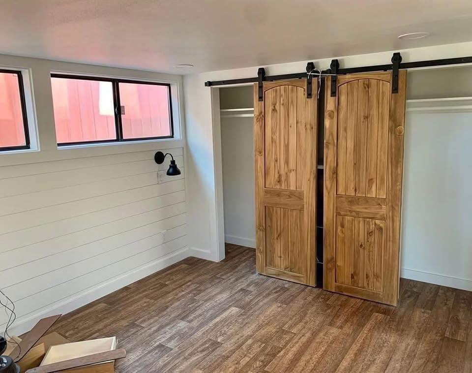 A cozy room with wooden sliding barn doors concealing a closet. The room features laminate wood flooring, white shiplap walls, and a black wall sconce.
