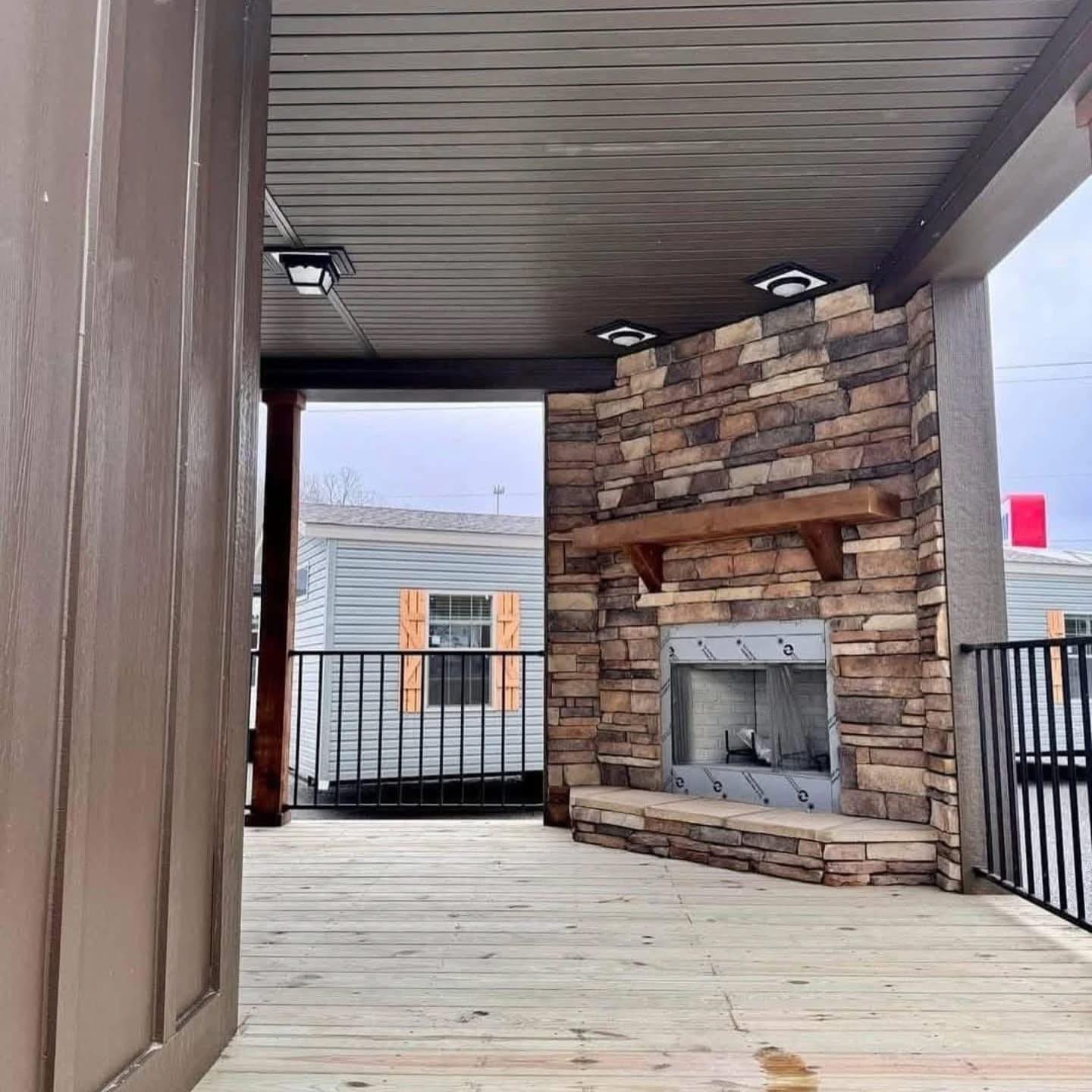 Covered wooden patio with stone fireplace and ceiling lights. Black railing surrounds the area. Cozy and inviting outdoor space with a modern design.