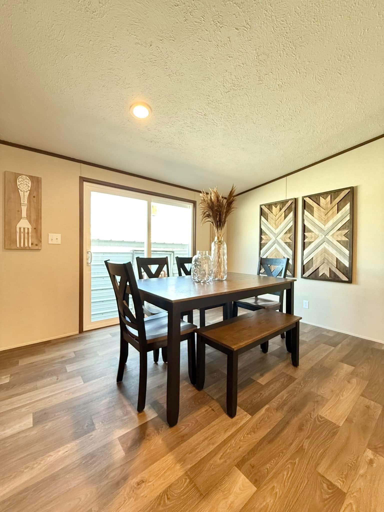 A cozy dining room features a wooden table with three chairs and a bench. Earth-toned geometric art and a vase with pampas grass add warmth.