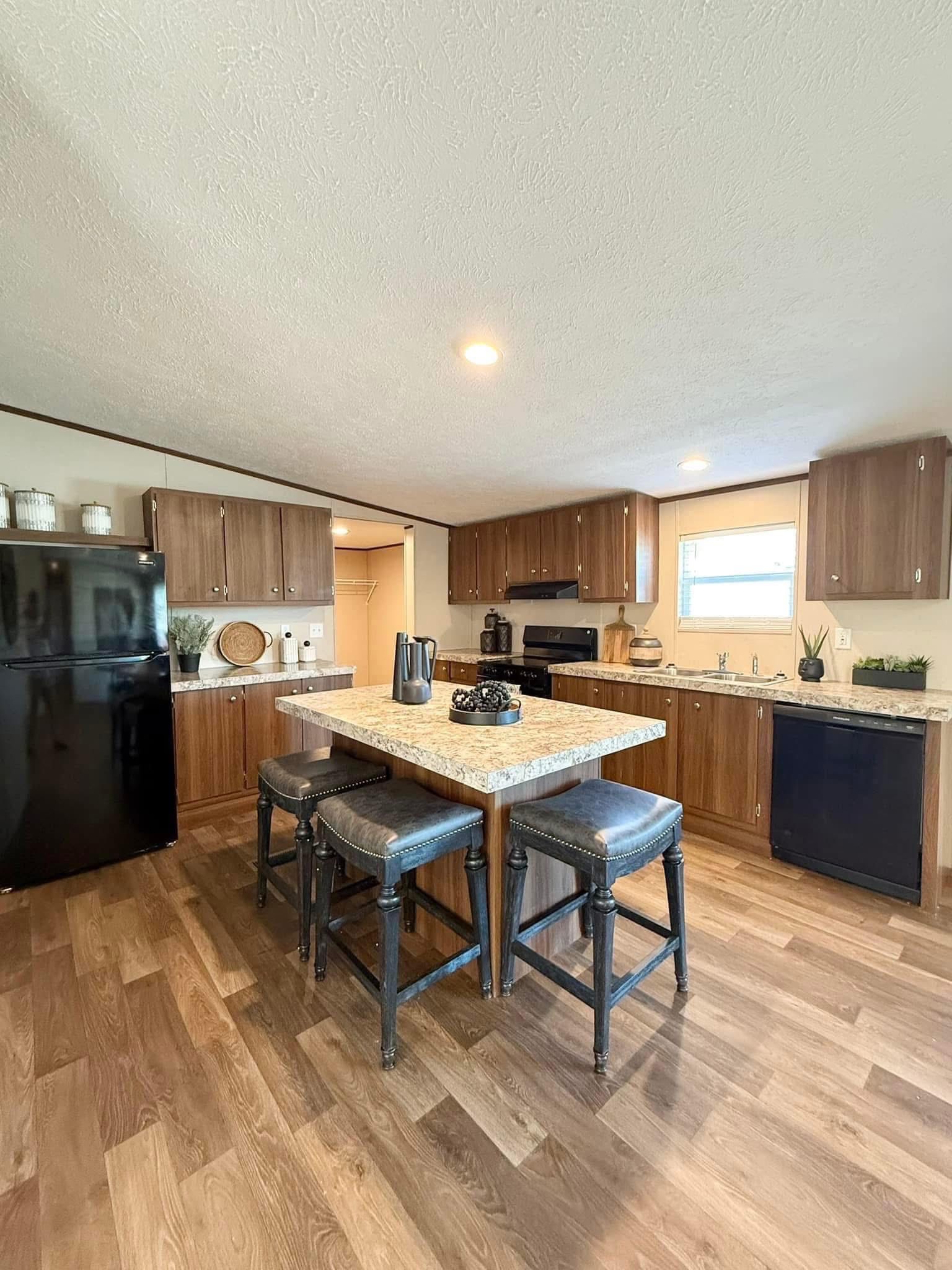 Modern kitchen with wood cabinets, sleek black appliances, and a central island featuring three cushioned stools. Warm tones create a cozy, inviting atmosphere.
