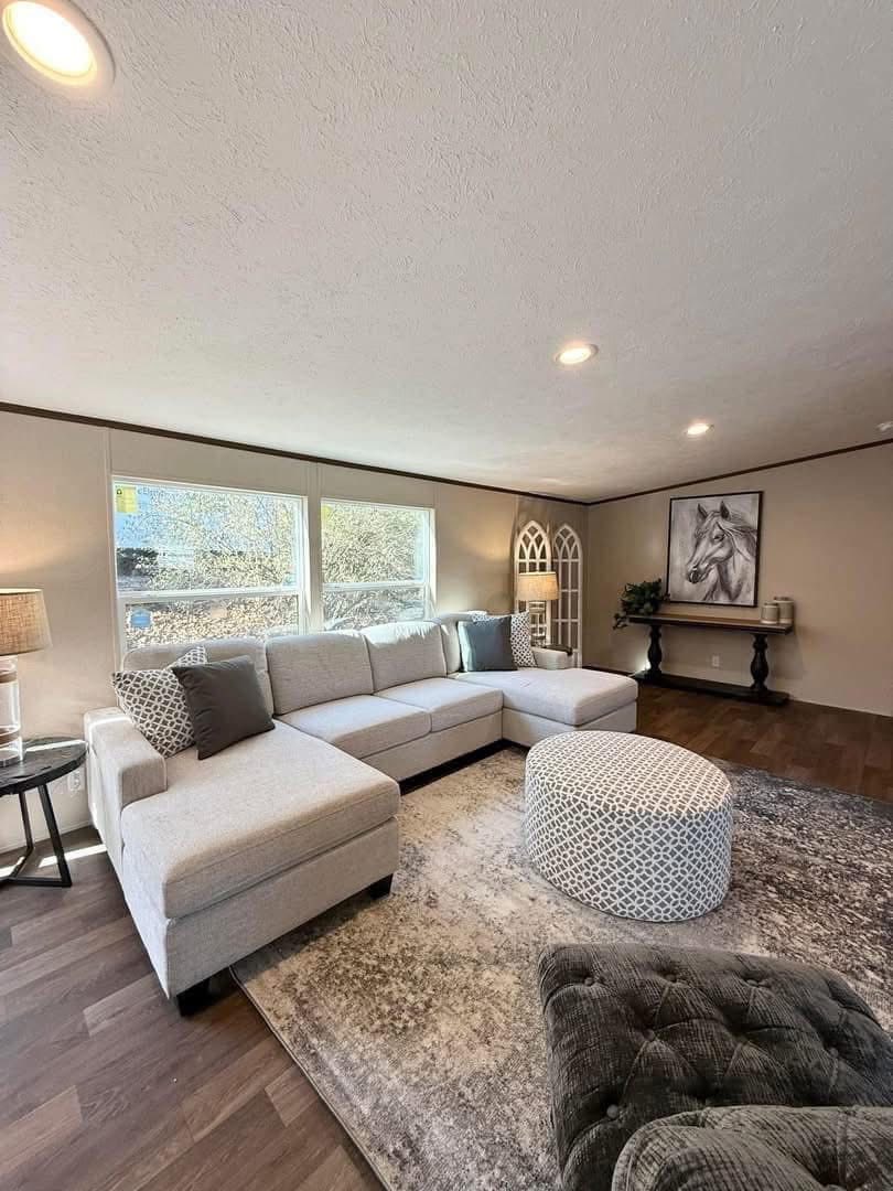 A cozy living room with a large beige sectional sofa, patterned cushions, and a round ottoman. Sunlight filters through windows, creating a warm, inviting atmosphere.