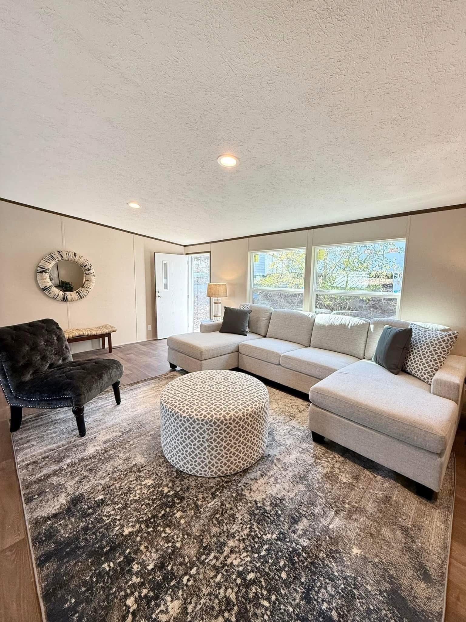 Modern living room with a large beige sectional sofa, patterned ottoman, and dark accent chair on a textured rug. Bright natural light streams through a window.