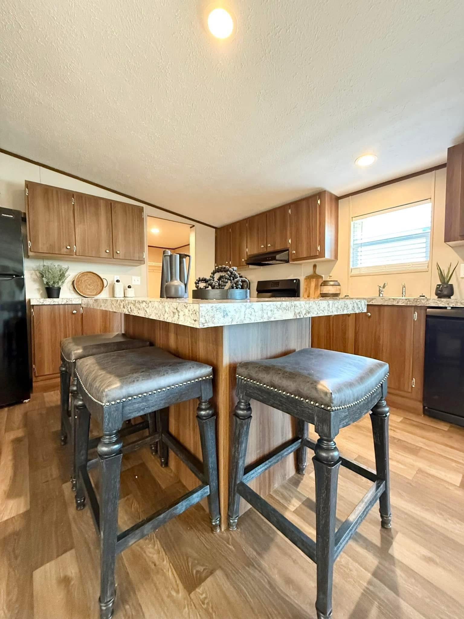 Modern kitchen with a granite-topped island and four dark leather stools. Wooden cabinets, black fridge, and sunlight through a window create a warm, inviting atmosphere.