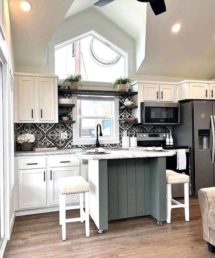 Bright living room with modern decor: beige sofa with green cushions, green armchair, brick fireplace, framed art, wood floors, and large window. Cozy ambiance.