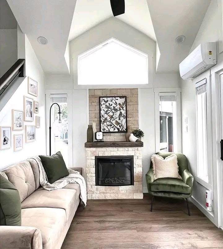 A cozy living room with high ceilings and neutral tones. A beige sofa and green armchair flank a brick fireplace. Artwork and decor create a serene atmosphere.