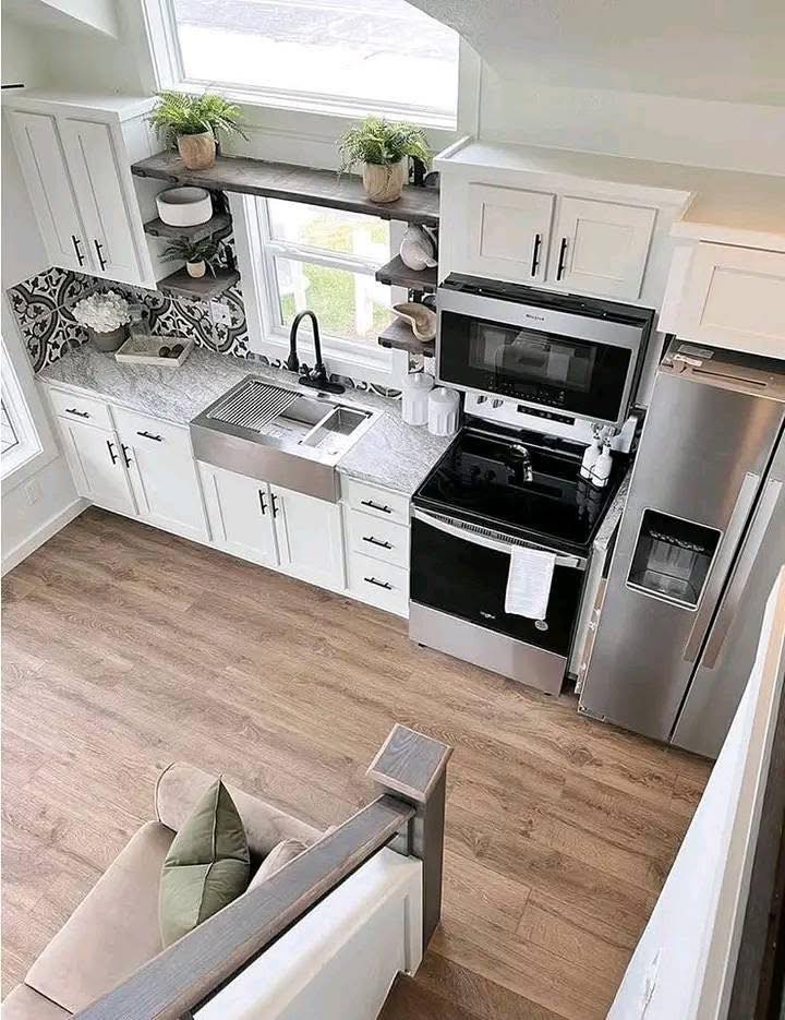 Bright kitchen with white cabinets, stainless steel appliances, and a farmhouse sink. Open shelves with plants. Cozy ambiance, light wood floor.