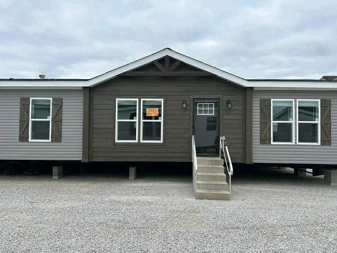A beige and brown modular home on gravel, with a small porch and stairs leading to the front door. The sky is overcast, creating a neutral mood.