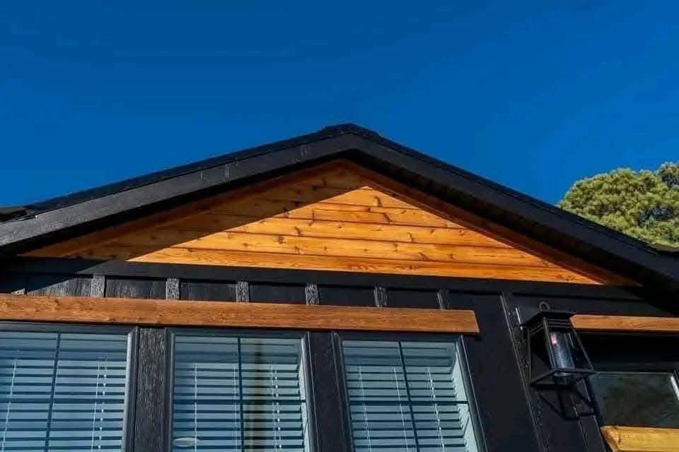 Close-up of a house's roof with black beams and warm wood paneling, beneath a bright blue sky. A black lantern is mounted on the wall.