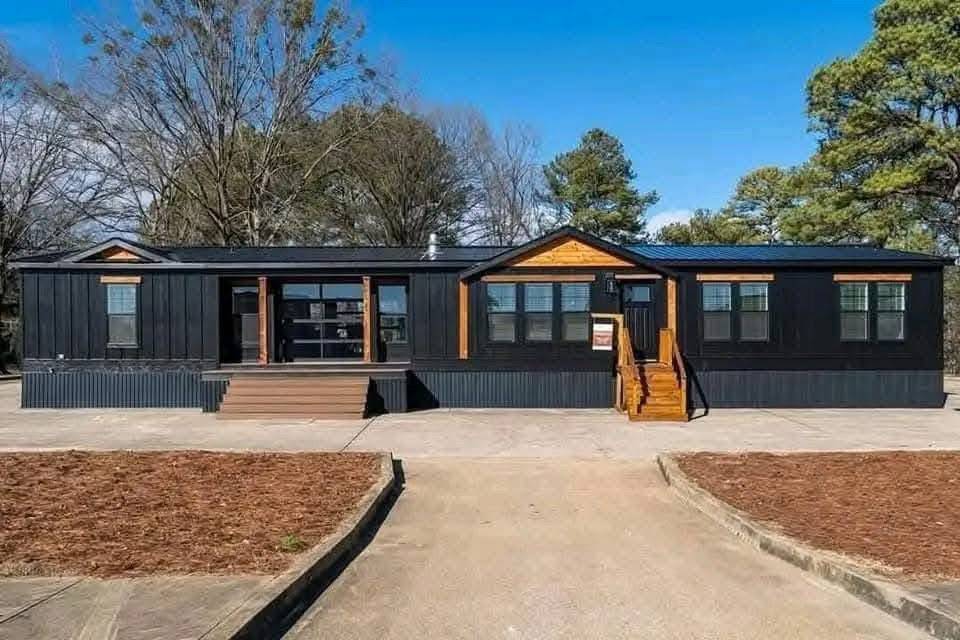 Modern black modular home with contrasting wooden accents, a central glass door, and steps. Surrounded by trees and clear blue sky. Peaceful setting.