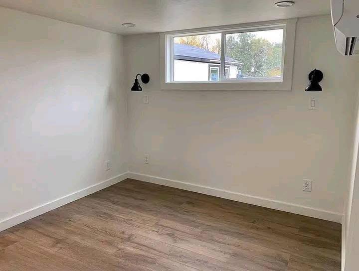 A small, minimalist room with white walls and wooden floor. Two black wall lamps flank a high, narrow window showing greenery outside. A calm, simple space.