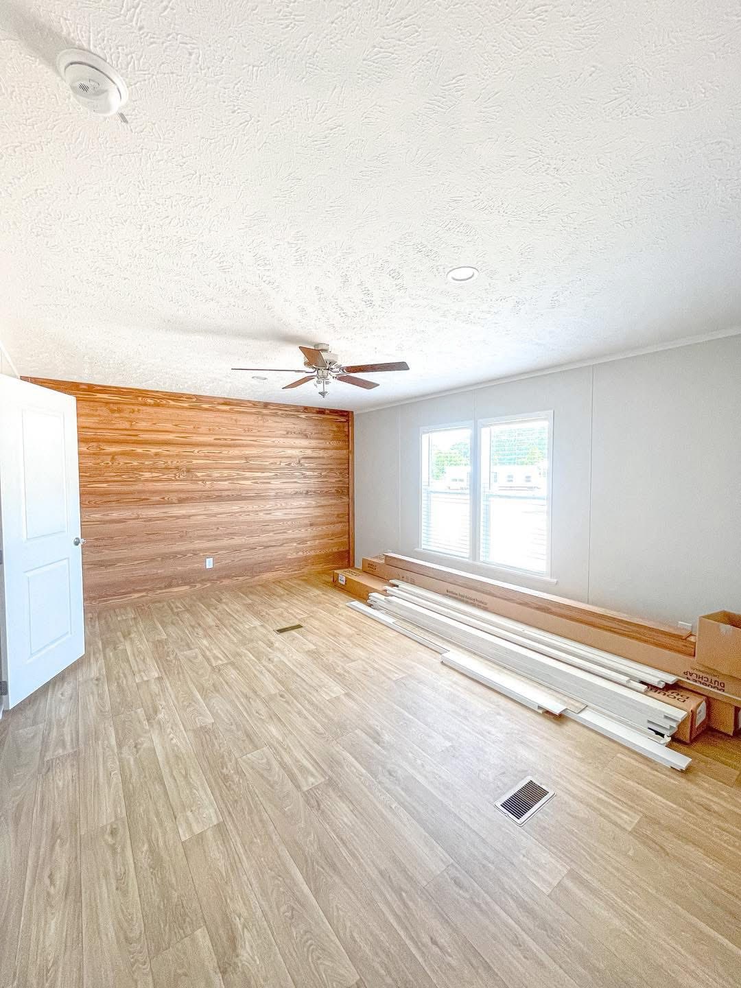 Empty room with light wood flooring, a wood accent wall, and a white ceiling fan. Sunlight streams through large windows onto stacked molding and boxes on the floor, conveying a sense of renovation.