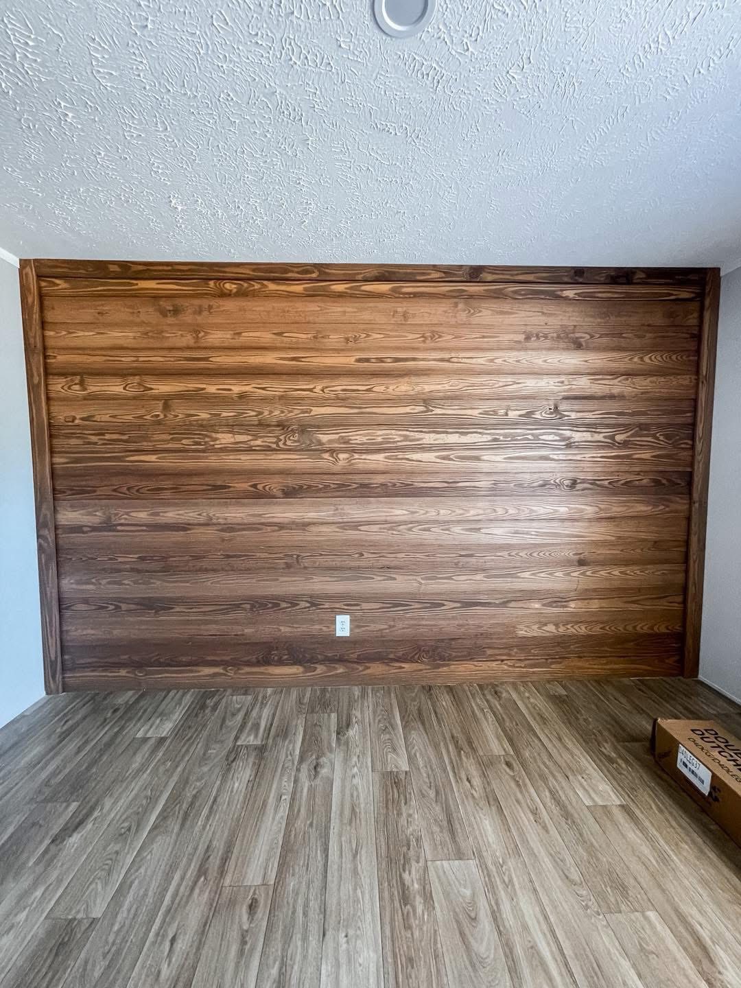 A room with a wooden accent wall and matching wood floorboards. The ceiling is textured white, and there's a small light fixture overhead. A cardboard box sits on the floor in the corner, creating a minimalist and calm atmosphere.