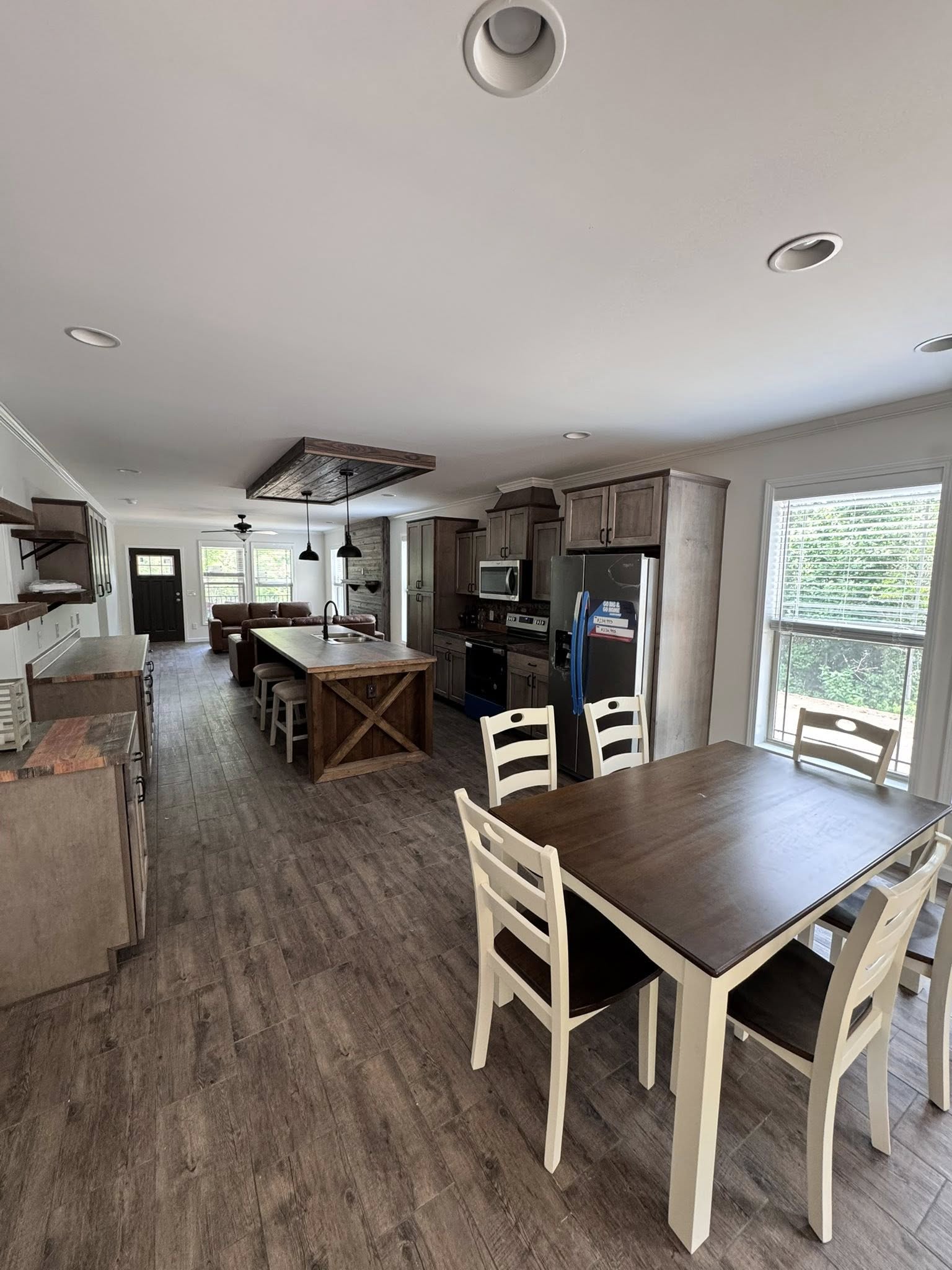 Rustic kitchen with wooden cabinets and island, stainless steel appliances, and a farmhouse sink. Living area visible with brown sofa and large windows. Cozy and inviting atmosphere.