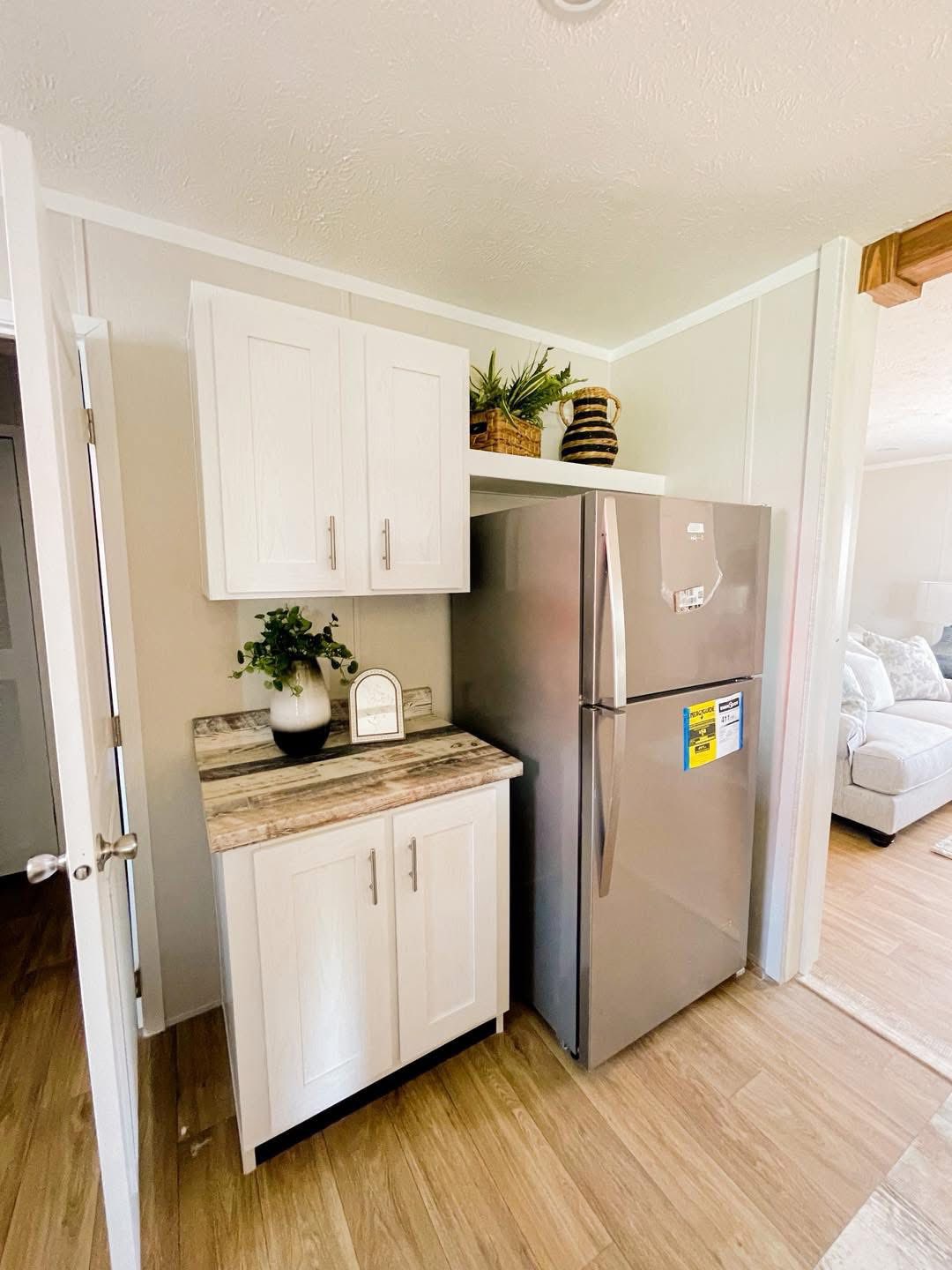 Compact kitchen nook with white cabinets, a granite countertop, and a stainless steel fridge. Decor includes plants in woven baskets on top of the fridge, adding a fresh, cozy vibe. Light wood flooring enhances the bright, airy atmosphere.