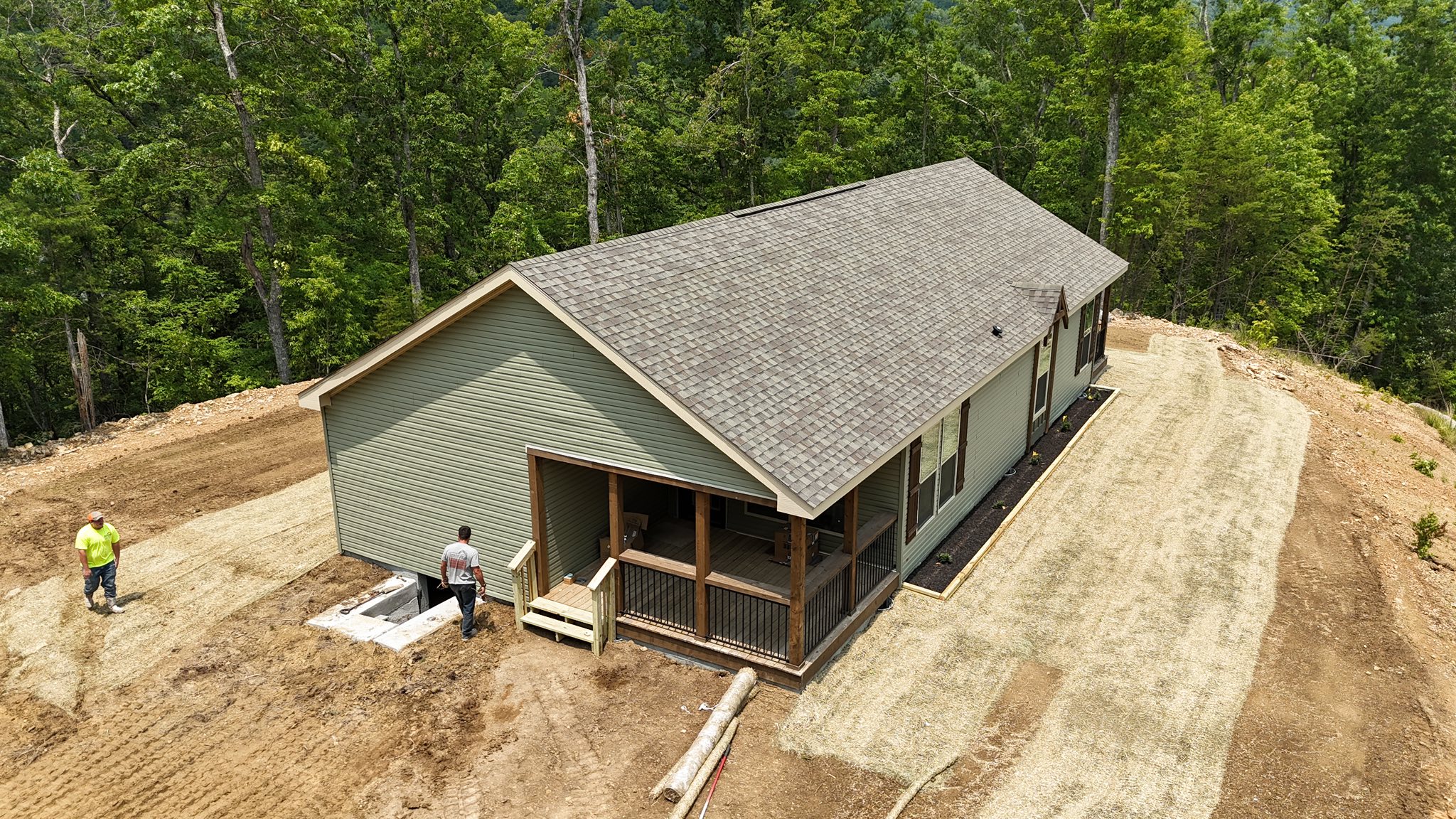 Single-story green house with a sloped roof, surrounded by a dirt yard and dense trees. Two people stand nearby, possibly engaged in construction work.