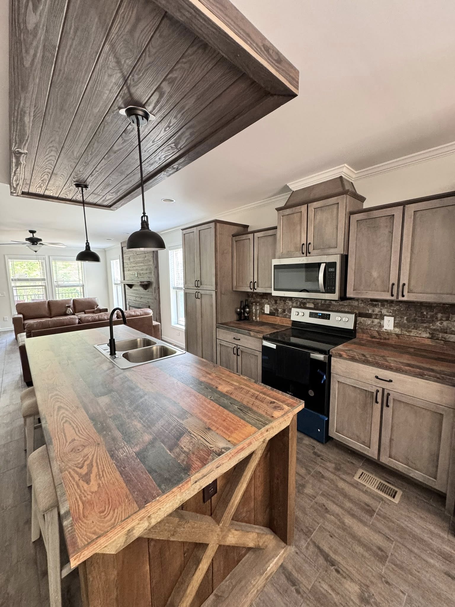 Rustic kitchen with wooden cabinets and island, stainless steel appliances, and a farmhouse sink. Living area visible with brown sofa and large windows. Cozy and inviting atmosphere.
